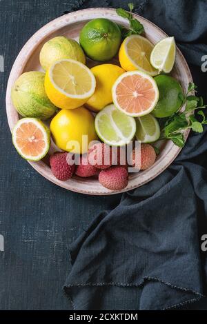 Variety of whole and sliced citrus fruits pink tiger lemon, lemon, lime, mint and exotic lichee on plate with textile over dark blue canvas textured b Stock Photo