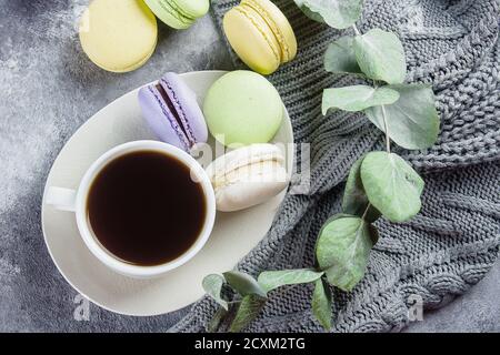 Cozy Morning Home Concept. Delicious colorful pastel macarons with cream and coffee cup, warm gray sweater on gray background. Top view Stock Photo