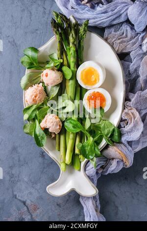 Cooked green asparagus with half boiled egg, salad, red caviar and salmon pate served on white fish shape plate with gauze textile over gray blue text Stock Photo