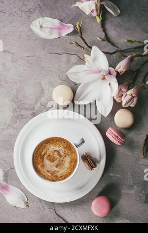 White cup of black coffee, served on white saucer with macaroons biscuits and magnolia flower blossom branch over gray texture background. Flat lay, s Stock Photo