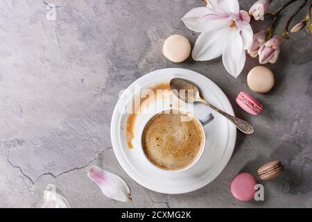 White cup of black coffee, served on white saucer with macaroons biscuits, spoon and magnolia flower blossom branch over gray texture background. Flat Stock Photo