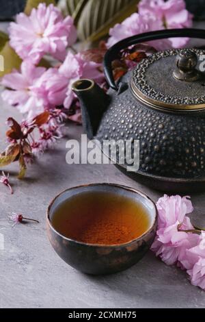 Black iron teapot and traditional ceramic cup of tea with blossom pink flowers cherry branch over gray texture background. Asian style. Stock Photo