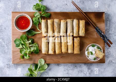 Fried spring rolls with red and white sauces, served on wood serving board with fresh green salad and wooden chopsticks over gray blue texture backgro Stock Photo