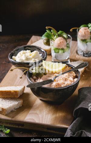 Black bowl of salmon pate with red caviar served with butter, sliced bread, capers, vintage knife, verrines and herbs on wooden serving board, textile Stock Photo