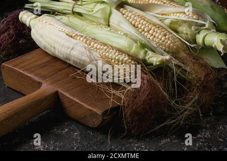 Young raw uncooked corn cobs in leaves on wooden chopping cutting board. Close up over dark brown concrete texture background. Stock Photo