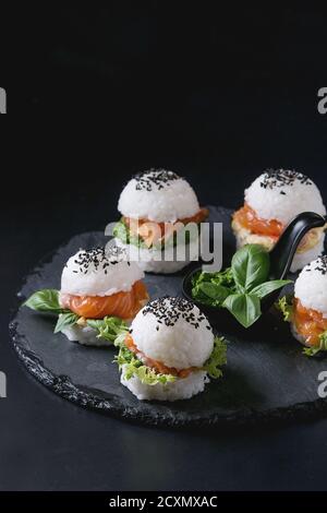 Mini rice sushi burgers with smoked salmon, green salad and sauces, black sesame served on slate stone board over black background. Modern healthy foo Stock Photo
