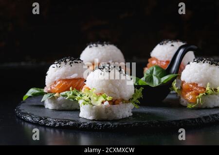 Mini rice sushi burgers with smoked salmon, green salad and sauces, black sesame served on slate stone board over black background. Modern healthy foo Stock Photo
