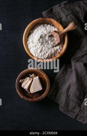 Fresh organic broken yeast cube, olive wood bowl of flour for baking homemade bread. With scoop, serving board, textile over black burned wooden backg Stock Photo