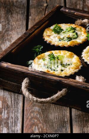 Baked homemade quiche pie in mini metal forms served with fresh greens in dark wood tray on old plank wooden background. Stock Photo