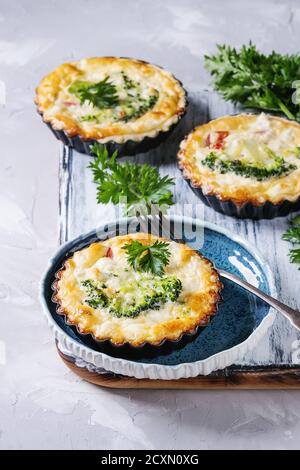 Baked homemade vegetable broccoli quiche pie in mini metal forms served with fresh greens, plate, fork on white serving board on gray concrete backgro Stock Photo