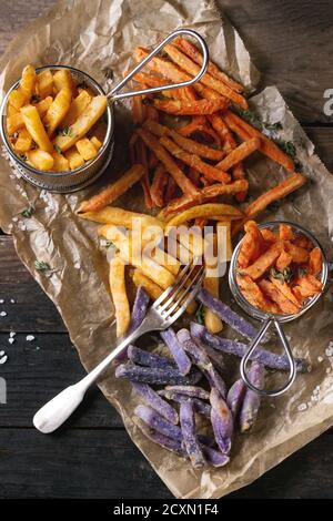 Variety of french fries traditional potatoes, purple potato, carrot served with salt, thyme and fork on baking paper over old wooden background. Top v Stock Photo