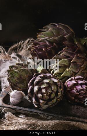 Uncooked whole organic wet purple artichokes with garlic in old rusty oven tray on textile sackcloth over dark wooden background. Rustic style. Close Stock Photo