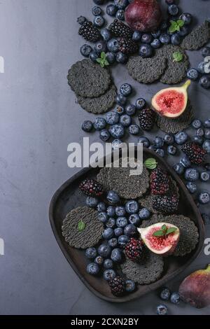 Variety of fresh berries blueberry, dewberry, red currant and figs with black charcoal crackers on wooden plate over dark metal background. Top view w Stock Photo