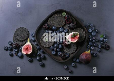 Variety of fresh berries blueberry, dewberry, red currant and figs with black charcoal crackers on wooden plate over dark metal background. Top view w Stock Photo
