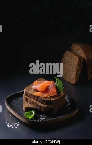 Stack of sliced homemade rye bread with smoked salmon, sea salt and fresh basil on wooden plate over dark black background. Stock Photo