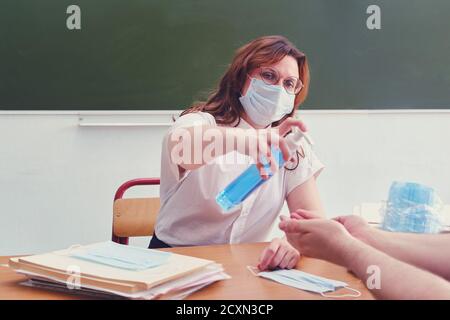 Woman school doctor treats his hands with antiseptic. Teacher in medical mask uses hand sanitizer, copy space. Classroom safety concept during lessons Stock Photo