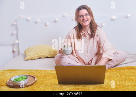 Happy woman studying online with laptop on home bed. Student remotely learns during isolation due to coronavirus Stock Photo