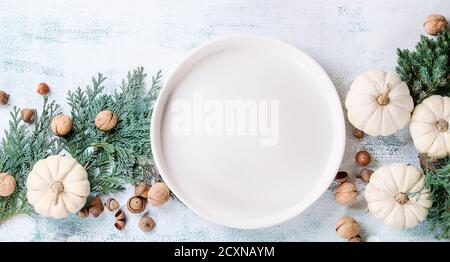 Holiday table decoration with white decorative pumpkins, thuja branches, walnuts, acorns and empty dinner plate over white wooden background. Top view Stock Photo