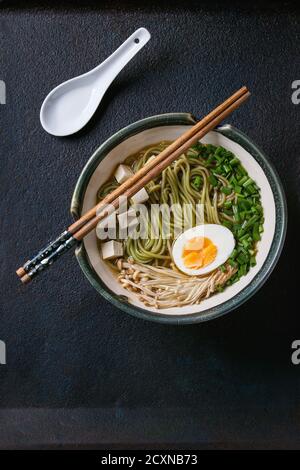Bowl of asian style soup with green tea soba noodles, egg, mushrooms, spring onion and tofu cheese, served with chopsticks and white spoon over black