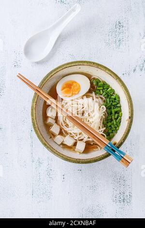 Ceramic bowl of asian style soup with noodles, half of egg, spring onion and tofu cheese, served with chopsticks and spoon over white wooden backgroun Stock Photo