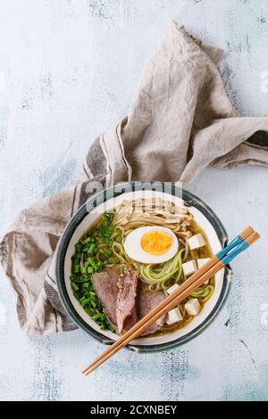 Bowl of asian style soup with green tea soba noodles, egg, mushrooms, beef, spring onion and tofu cheese, served with chopsticks and textile over whit Stock Photo