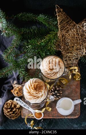 Pumpkin spicy latte  with whipped cream and cinnamon in two glasses standing on clay board with textile and Christmas decoration fir tree other dark b Stock Photo