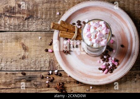Glass of cafe latte with pink marshmallow standing on ceramic plate with spices, coffee beans and pink sugar over wooden texture background. Top view Stock Photo