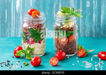 Salads with quinoa, couscous, spinach, radish, tomatoes and zucchini in glass mason jars, standing with fresh vegetables over bright turquoise wooden Stock Photo