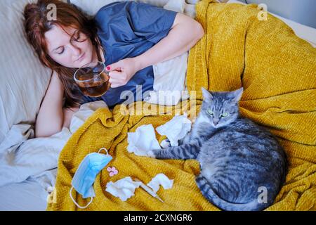 Cat in bed of a sick woman drinking tea. Woman with a coronavirus drinks tea while lying on a bed next to a cat