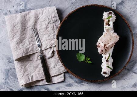 Food plating french dessert chocolate meringue with fresh mint, whipped cream and coffee beans served with fork and textile napkin on black plate over Stock Photo