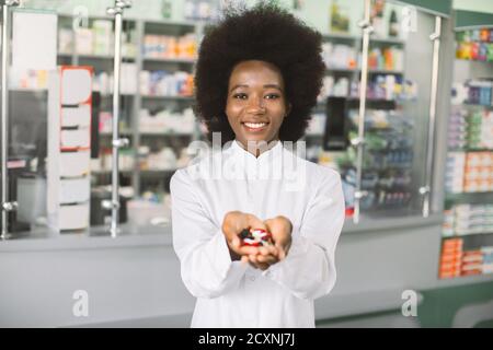 Young smiling African-american female pharmacist, holding a handful of medicine pills and colorful tablets in the hands, offering to camera. Drug Stock Photo