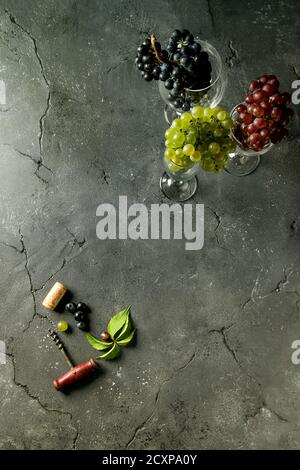Variety of three type fresh ripe grapes dark blue, red and green in different laying wine glasses with old corkscrew and green leaves over black textu Stock Photo