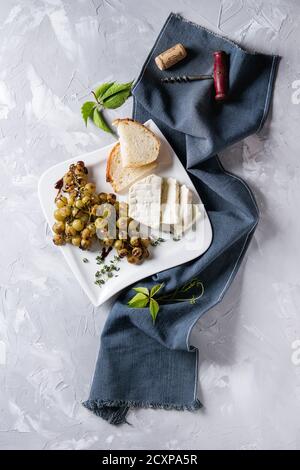 White plate with sliced camembert cheese and baked bunch of green grapes served with bread, wine glass, corkscrew, green leaves, fork on blue textile Stock Photo