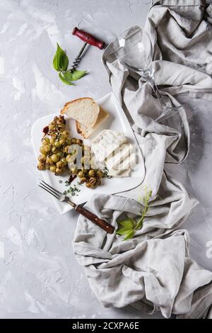 White plate with sliced camembert cheese and baked bunch of green grapes served with bread, wine glass, corkscrew, green leaves, fork on textile over Stock Photo