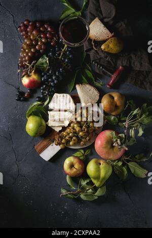 Serving board with sliced camembert cheese and baked bunch of green grapes served with bread, glass of red wine, corkscrew, apples, pears, leaves over Stock Photo
