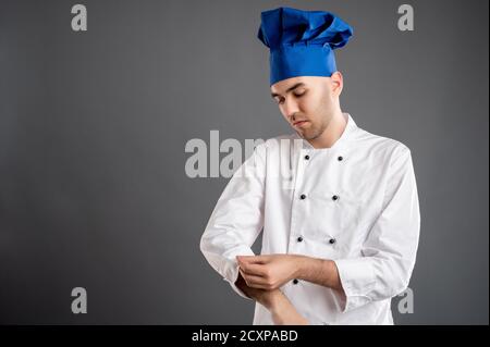 Portrait of young male dressed in a white chef suit arranges his uniform posing on a gray background with copy space advertising area