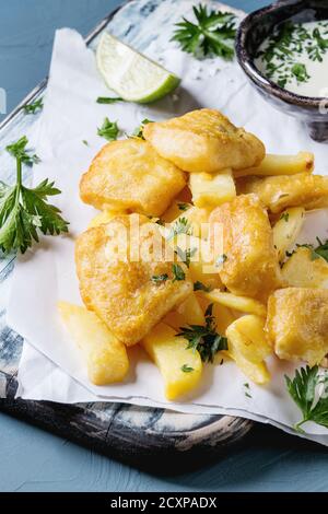 Traditional british fast food fish and chips. Served with white cheese sauce, lime, parsley, on white paper and black serving board over blue concrete Stock Photo