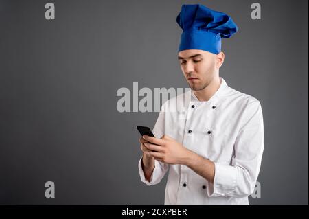 Portrait of young male dressed in a white chef suit with blonde hair, reading and sending text messages on cellphone posing on a gray background. Stock Photo