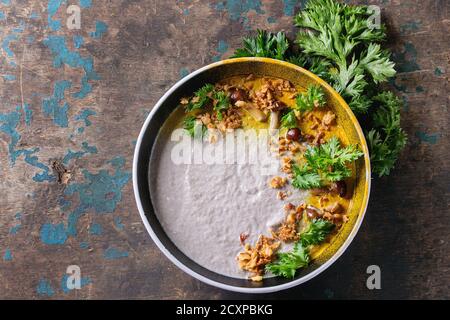 Mushroom cream soup in black ceramic bowl served with turmeric powder, forest mushrooms, greens parsley, fried onion over old dark wooden background. Stock Photo