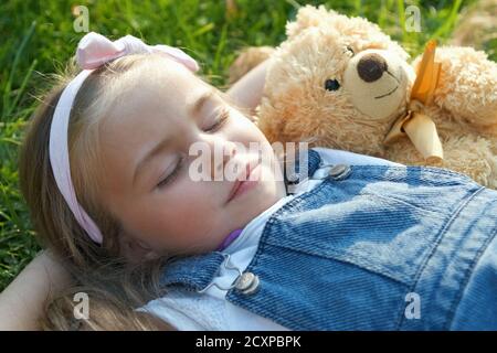 Pretty little child girl with closed eyes laying down with her teddy bear toy on blanket on green grass in summer taking a nap. Stock Photo