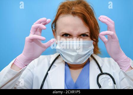 Woman doctor in a blue uniform is angry, copy space. Portrait of a readhead nurse on a blue background, close-up. Stay home. Stock Photo