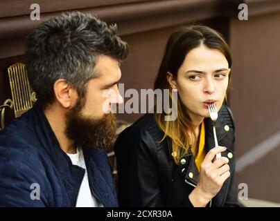 Woman and man with surprised faces have date at cafe. Urban cafeteria concept. Girl looks at bearded guy holding fork in mouth on brown terrace background. Couple in love takes a bite during break Stock Photo