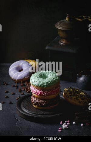 Variety of colorful glazed donuts with chopped chocolate, pink sugar, coffee beans vintage coffee grinder on black texture background. Dark rustic sti Stock Photo
