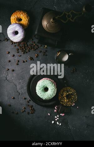 Variety of colorful glazed donuts with chopped chocolate, pink sugar, coffee beans vintage coffee grinder on black texture background. Dark rustic sti Stock Photo