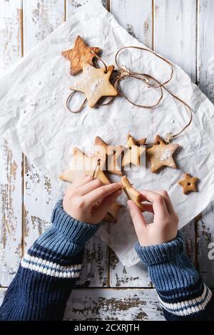 Child hands make garland of homemade shortbread star shape sugar cookies different size on thread on baking paper over white wooden plank table. Chris Stock Photo