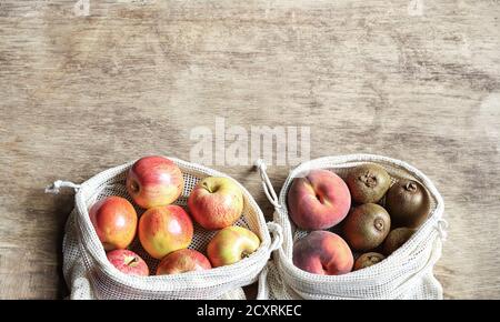 Organic fresh fruits - apples, kiwi, peaches in reusable eco friendly natural bags on the wooden background. Package-free food shopping. Stock Photo