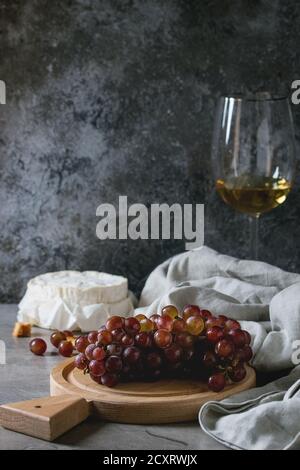 Bunch of red grapes, camembert cheese, croutons and glass of white wine served on wooden serving board over gray kitchen table. Rustic style. Stock Photo