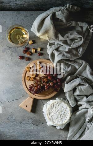 Bunch of red grapes, camembert cheese, croutons and glass of white wine served on wooden serving board over gray kitchen table with textile linen. Rus Stock Photo