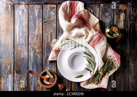 Mediterranean style table with white empty plate, kitchen towel, olive wood branch, olive oil, garlic, sea salt, pepper over old plank background. Top Stock Photo