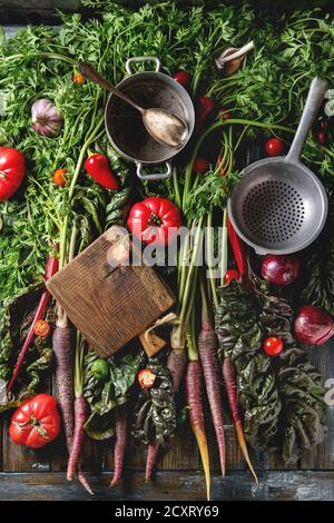 Raw organic purple carrot with variety of vegetables tomatoes, pepper, chard, garlic, onion, empty pan, colander, cutting board on haulm over old wood Stock Photo